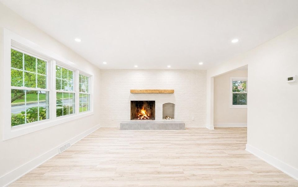 Living room with front windows, painted brick fireplace and wide-plank flooring.