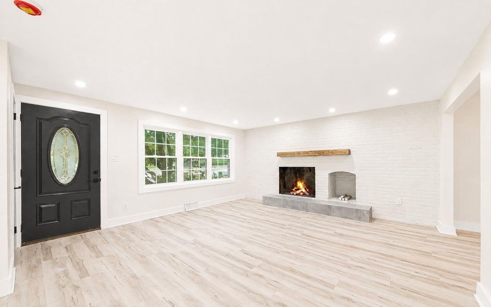 Living room after the renovation: black front door with oval glass, new wide-plank flooring, recessed lighting and the original brick fireplace painted white.