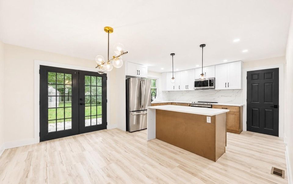 Dining area and kitchen: French doors to the rear patio, a brass-and-glass chandelier, stainless refrigerator, white upper cabinets, beige lower cabinets and a quartz waterfall island.