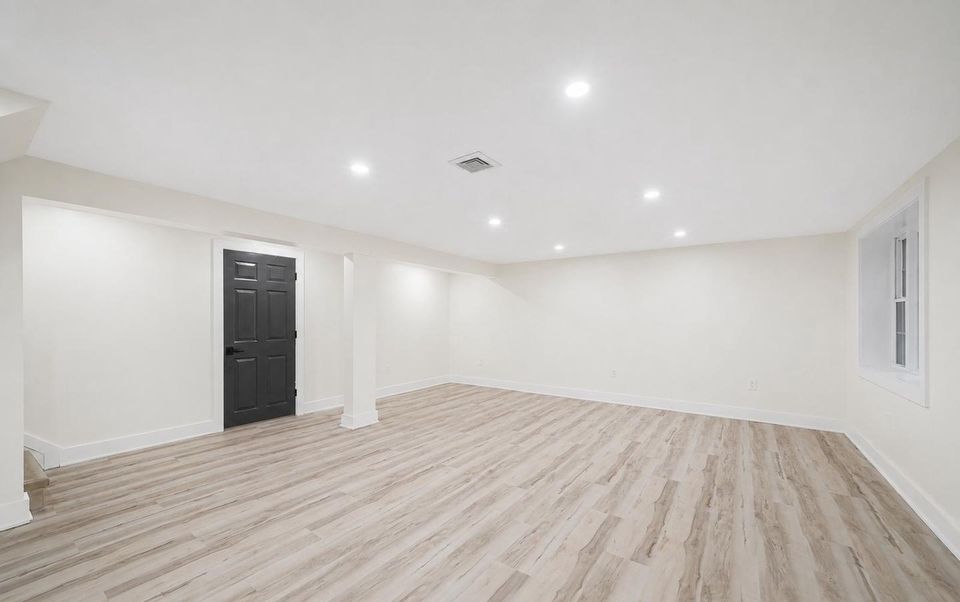 After: the finished lower-level recreation room with wide-plank flooring, white walls, recessed lighting and the stair to the main level.