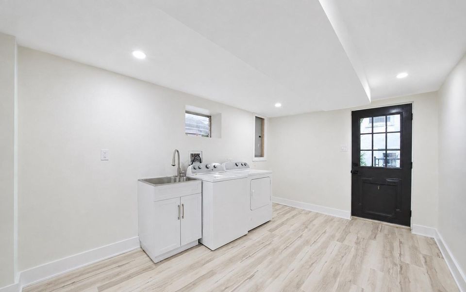 After: the finished laundry with white walls, recessed lighting, a new utility-sink cabinet, washer, dryer and a door to the outside.