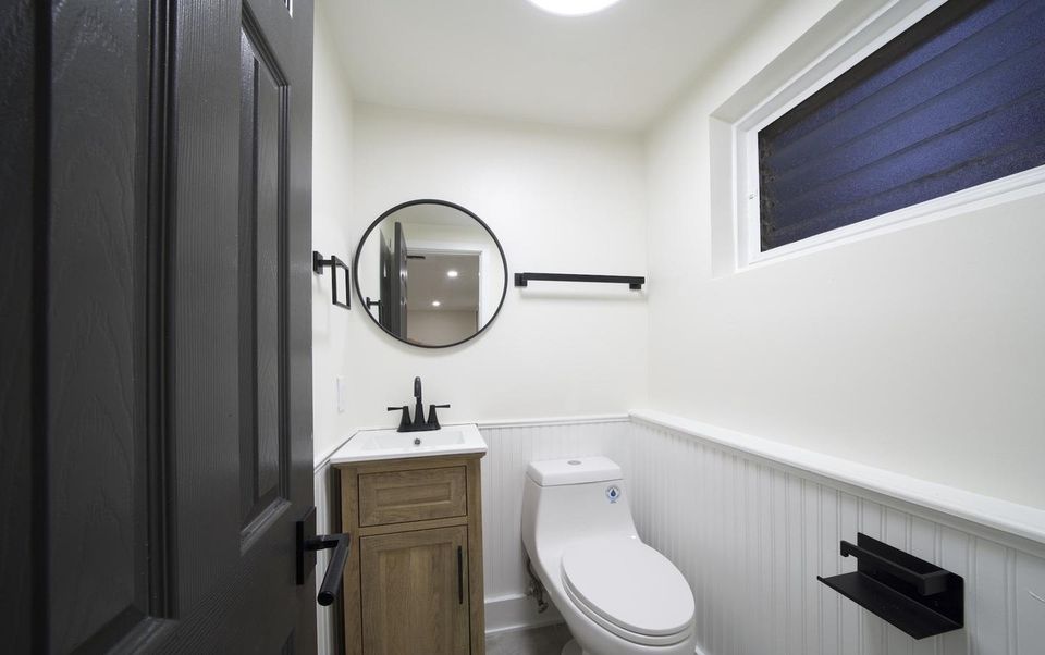 Powder room with beadboard wainscoting, wood vanity, round mirror and black fixtures.